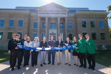 A group of people stands in front of the University of Kentucky Agricultural Experiment Station building during a ribbon-cutting ceremony. The historic brick building features a columned entrance and a sign reading “AGRICULTURAL EXPERIMENT STATION." 