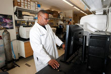 Person wearing a white lab coat, works at a computer beside research equipment in a laboratory.