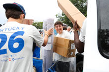 A smiling student in a clear rain poncho carries boxes as volunteers unload move-in supplies from a van.