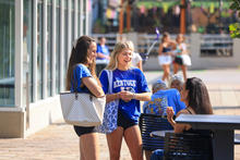 Two smiling University of Kentucky students chat with seated attendees outside a campus building on a sunny day.