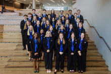 A large group of students wearing matching professional attire and blue scarves pose on tiered wooden steps inside a campus building.