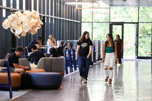 Two students walk together through a bright campus lounge as others study and socialize nearby.