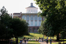 A large academic building stands at the end of a tree-lined campus walkway. Students walk along the path and gather on grassy areas beneath the trees on a sunny day.