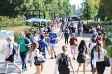 Students walk along a busy campus sidewalk on a sunny day during the start of the fall semester. People carrying backpacks travel in both directions along a tree-lined pedestrian pathway.