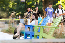 Person sitting in a colorful Adirondack chair outdoors, using a laptop and holding a mug while students walk nearby.