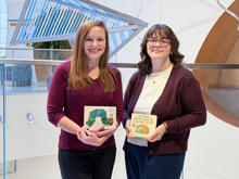 Two people stand indoors holding children’s books, with a large blue-lit art installation behind them.