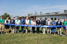 A group of people standing outdoors in front of a large modern industrial or educational facility, holding a blue ceremonial ribbon and scissors during a ribbon-cutting event. The building features metal siding and rooftop ventilation equipment.