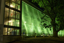Exterior view of a multi-story building illuminated with bright green lighting at night. A large translucent green facade stretches along the structure, while an adjacent glass stairwell reveals interior landings and railings.
