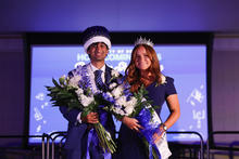 Two people wearing Homecoming crowns and sashes pose together while holding bouquets of flowers.