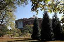 People walk along a tree-lined path outside a red brick campus building, framed by green and yellow autumn leaves under a clear blue sky.