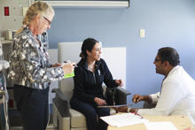 Three people gather in a clinical exam room, with one person seated and speaking with another seated person in a white coat while a third person stands nearby.