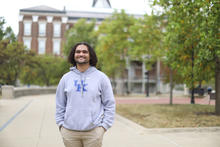 Person wearing a gray UK hoodie stands on a campus walkway.