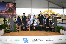 A group of people wearing hard hats pose behind a ceremonial dirt pile with shovels at a groundbreaking event under a large white tent. A banner along the front displays the UK HealthCare logo and the names Perkins&Will and Walsh.