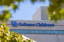 Exterior of the Golisano Children’s building featuring the blue UK logo and “Golisano Children’s” sign, framed by green tree leaves against a blue sky.