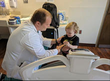 An adult wearing a white coat sits beside a hospital bed and holds a small medical instrument near a child seated in the bed, with medical supplies and a sink visible in the room.