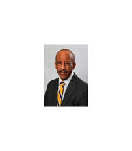 Portrait of a man wearing glasses, a dark suit, white shirt and blue-and-gold striped tie against a gray background.