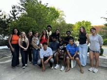 A group of young adults pose outdoors, standing and sitting around a bench with leafy trees behind them.