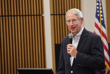 Person speaking into a handheld microphone at a podium in an indoor event space. An American flag is visible in the background, along with wood-paneled walls and presentation equipment.