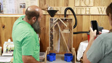 A person pours material from a cup into a funnel atop a transparent, wood-framed seed-sorting machine during a workshop demonstration.
