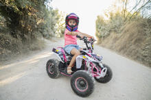 Child wearing a pink helmet and pink shirt sits on a small pink and black ATV on a dirt road lined with trees and dry grass.