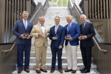 Five alumni award recipients stand on a marble staircase holding medals and plaques during a recognition ceremony.