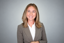 A smiling woman with shoulder-length blonde hair wears a gray blazer and white shirt against a soft gray background.