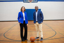  Two people stand on a gym basketball court beneath a hoop, with a basketball on the floor between them.