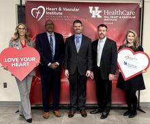 Five people stand in front of a Heart & Vascular Institute and UK HealthCare Gill Heart & Vascular Institute Network backdrop. Two are holding large heart-shaped signs, one reading “Love Your Heart.”