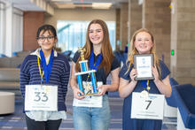 Three Big Blue Bee participants smile while wearing medals. The student in the center holds a champion trophy, and the student on the right holds a framed runner-up award.