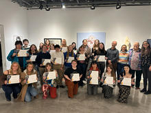 Group photo in an art gallery; people holding certificates, artworks displayed on walls.