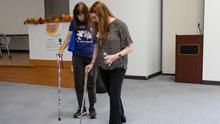 Person using forearm crutches walks across an indoor room while another person walks beside them, appearing to guide or assist with movement practice.