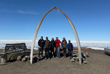 Six people stand beneath a large arch formed by two whale bones on a dark shoreline, with wooden boats and a vast expanse of sea ice beneath a clear blue sky in the background.