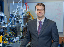 A person wearing a dark suit and patterned tie stands beside laboratory equipment, including stainless-steel scientific instruments, tubing and electronic components. A whiteboard with faint technical diagrams is visible in the background.