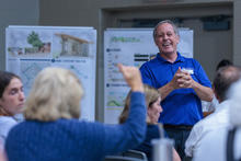 A person in a blue polo shirt stands at the front of a community meeting or public workshop, speaking to attendees seated in the foreground. Large presentation boards displaying renderings, maps, and planning diagrams are positioned behind the speaker. 