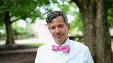 Outdoor portrait of an adult wearing a white shirt and pink bow tie, standing under leafy trees. The person faces the camera with a slight smile, with a softly blurred campus-like background.