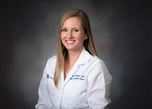 Woman wearing a white medical lab coat with embroidered text on the chest, posed against a dark gray studio background.