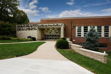 Brick academic building with a covered entrance, walkway and landscaping on a sunny day.