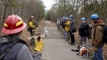 Field leader on the left speaking to several students on the right all wearing heavy fire gear and helmets 