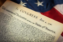 A close-up of the Declaration of Independence document dated July 4, 1776. The document is placed on an American flag, showing red and white stripes and part of a blue field with white stars in the background.