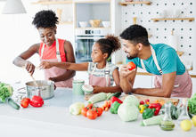 A family of three wearing aprons cooks together in a bright kitchen, with a woman stirring a pot on the stove.