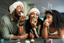 Three people wearing festive holiday hats lean over a kitchen counter, smiling and laughing while eating decorated cookies together, with sprinkles and baking supplies visible on the counter.
