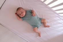 A baby lies on their back in a crib on a fitted white sheet, wearing a light blue polka-dot onesie.