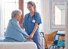 Health care professional in blue scrubs holding the hand of an older patient seated on a bed.