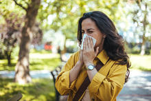 A woman wearing a yellow jacket sneezes into a tissue while standing outdoors near trees and greenery in spring.
