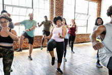 A group of adults participate in a high-energy group fitness or dance workout class in a bright studio with large windows and brick walls, lifting their knees and moving in sync while following an instructor at the front.