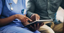 Health care worker in blue scrubs and a person in a gray-green shirt review information together on a tablet.