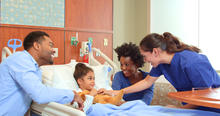 A child lies in a hospital bed while two adults sit nearby, and a health care provider stands at the bedside adjusting a blanket or comfort item. The room includes medical equipment and bedside furniture.