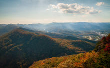 A panoramic view of forested Appalachian mountains covered in fall foliage, with a valley and community visible in the distance.