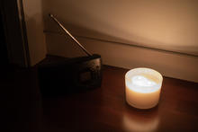 A small black radio with an extended antenna sits on a wooden surface next to a lit white candle, casting a warm glow against a beige wall.