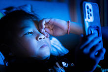 A child lies in bed in a dark room, holding a smartphone close to their face. Blue light from the phone illuminates the child's face and hand while the rest of the room remains dimly lit.